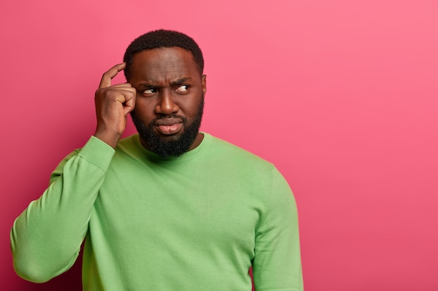 Foto grátis homem negro confuso e inconsciente coça a cabeça, franze a testa enquanto olha para o lado, sente dúvida ou hesitação ao tomar uma decisão, usa um macacão verde, isolado na parede rosa do estúdio