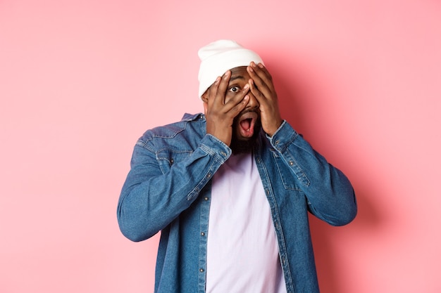 Homem negro curioso cobrindo os olhos, mas espiando por entre os dedos, olhando para a câmera espantado, usando um gorro hipster contra um fundo rosa