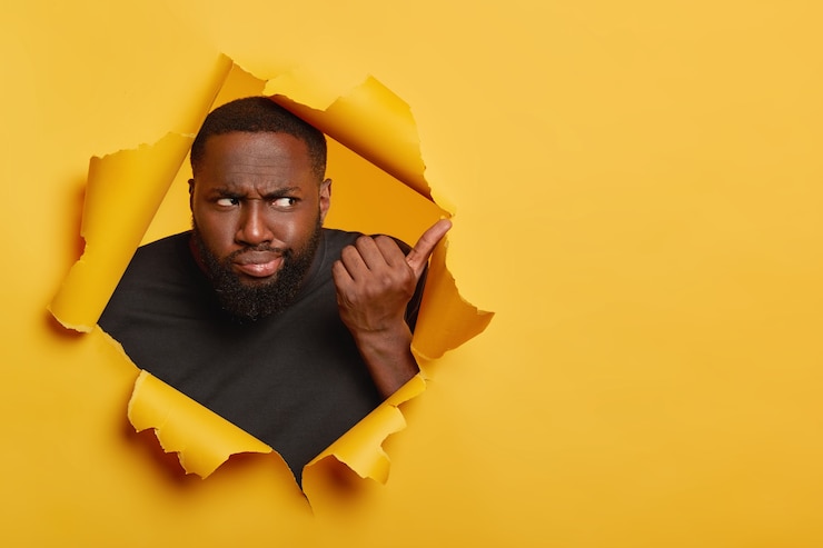 Foto grátis homem negro frustrado e infeliz com um sorriso afetado, aponta para longe com um olhar desconfiado e nada impressionado, posa em um fundo de papel amarelo rasgado