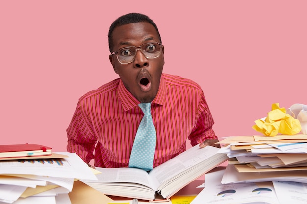 Foto grátis homem negro horrorizado e cômico segurando um livro grosso aberto, olhando em estupor, trabalhando em um papel do curso, tem uma pilha de papéis na mesa