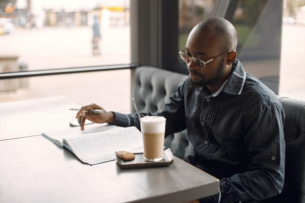 Homem negro sentado em um café e bebendo um café com leite. Homem usando óculos escuros e camisa cinza