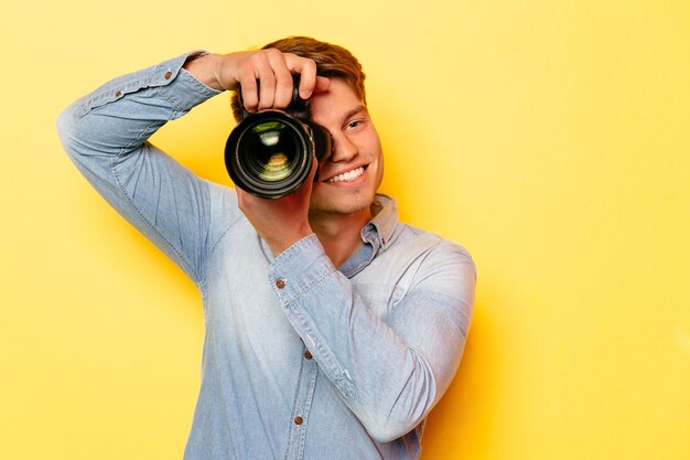 Homem novo alegre com câmera profissional, tomando uma foto. Sobre fundo amarelo.