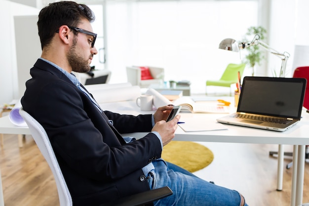 Homem novo considerável que usa seu telefone móvel no escritório.