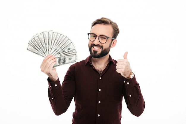 Homem novo de sorriso que mantém o dinheiro que mostra os polegares.