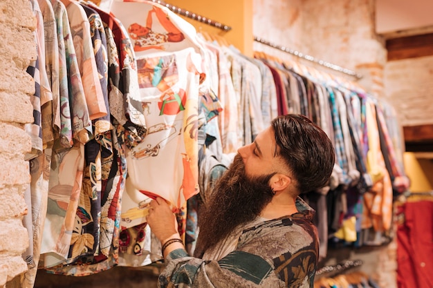 Homem novo farpado que escolhe a camisa que pendura no trilho na loja