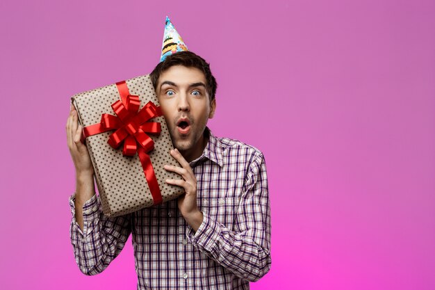 Homem novo feliz que guarda o presente de aniversário na caixa sobre a parede roxa.