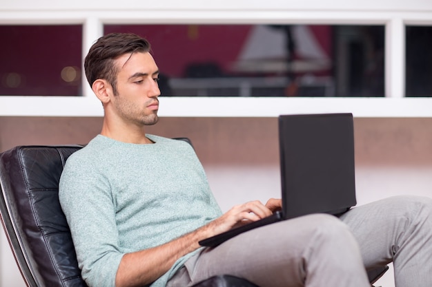 Homem novo sério que trabalha no portátil na cadeira