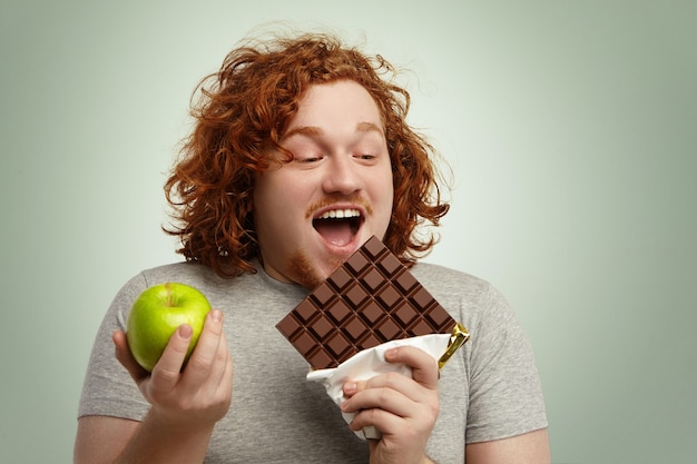 Homem obeso alegre abrindo amplamente a boca, segurando uma grande barra de chocolate em uma mão e maçã verde na outra