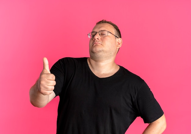 Foto grátis homem obeso de óculos, vestindo camiseta preta, parecendo confiante mostrando os polegares para cima sobre rosa