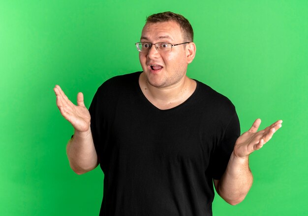 Homem obeso de óculos vestindo camiseta preta parecendo confuso e incerto estendendo os braços para os lados sem resposta sobre o verde