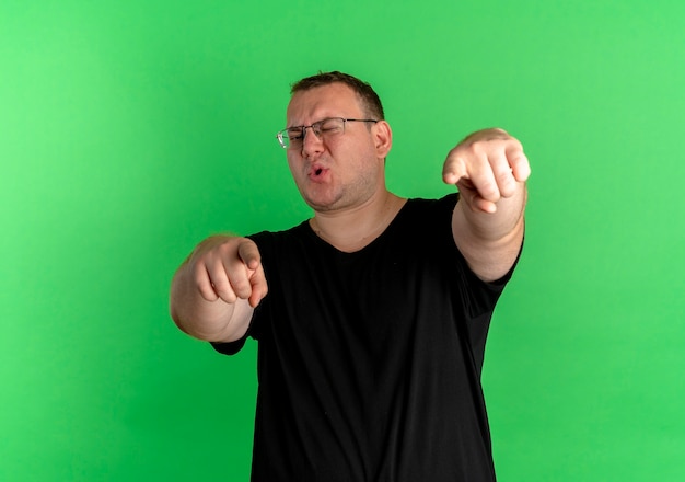 Foto grátis homem obeso de óculos, vestindo uma camiseta preta, parecendo confiante, apontando com o dedo indicador em pé sobre a parede verde