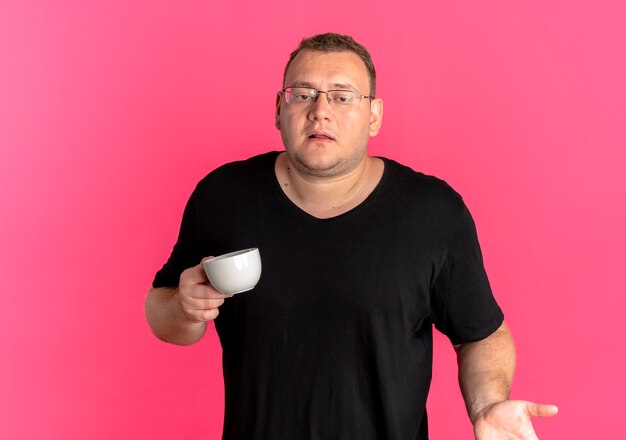 Homem obeso de óculos, vestindo uma camiseta preta segurando uma xícara de café, encolhendo os ombros e parecendo confuso em pé sobre uma parede rosa