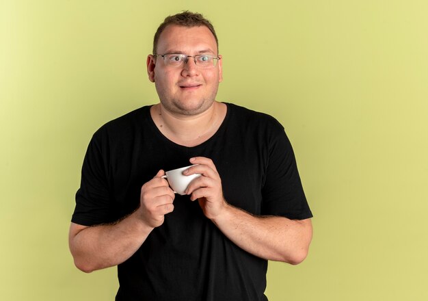 Homem obeso de óculos, vestindo uma camiseta preta segurando uma xícara de café, olhando de lado, emocionado e feliz em pé sobre uma parede de luz