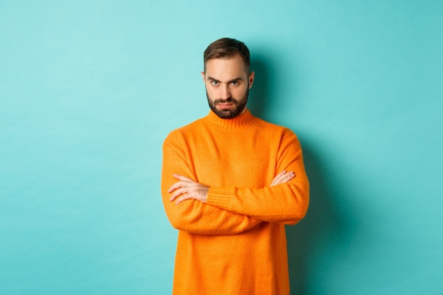 Homem ofendido, olhando com raiva para você, cruza os braços no peito e encara com raiva, em pé com um suéter laranja contra a parede turquesa.