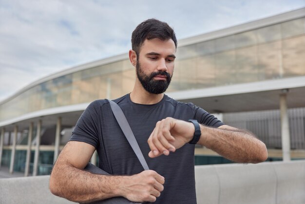 homem olha para smartwatch verifica tempo ou calorias queimadas após treino carrega karemat enrolado pratica ioga ao ar livre usa camiseta preta casual poses ao ar livre. Esporte na cidade