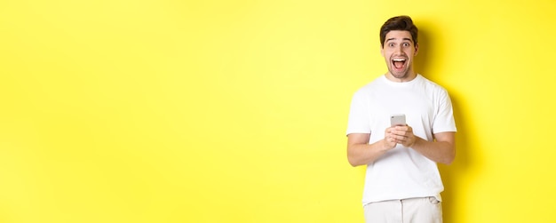 Foto grátis homem olhando espantado e feliz depois de ler algo no telefone móvel sobre fundo amarelo