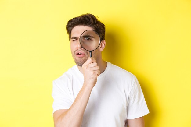 Homem olhando pela lupa com olhar sério e pensativo, procurando ou investigando, em pé sobre fundo amarelo.