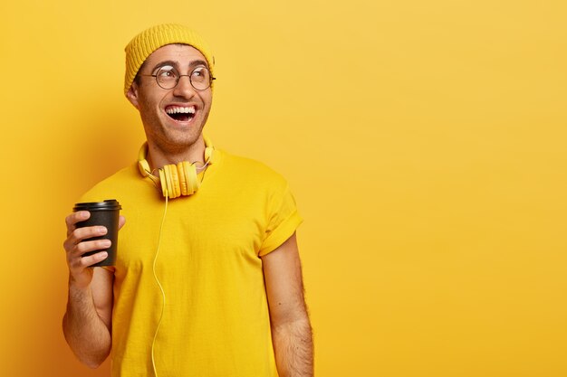 Homem otimista de óculos ri enquanto passa o tempo com os amigos durante a pausa para o café, segura um copo descartável, desvia o olhar