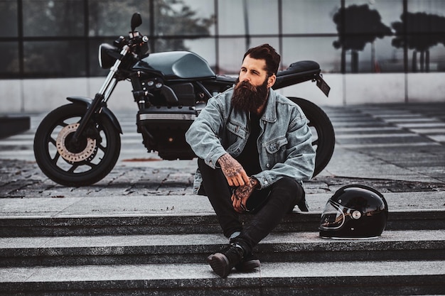 Foto grátis homem pensativo brutal de jaqueta jeans está sentado nas escadas perto de sua moto perto do prédio de vidro.