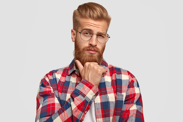 Foto grátis homem pensativo com a barba por fazer segura o queixo, olha pensativo diretamente para a câmera, pensa em algo importante, vestido com uma elegante camisa xadrez