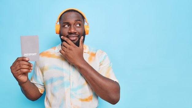 Homem pensativo de barba negra segura o queixo olhando para longe segura passaporte que contém informações básicas de identificação ouve música por meio de fones de ouvido sem fio isolados sobre a parede azul