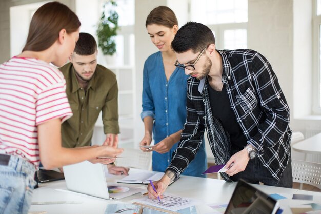 Homem pensativo de camisa e óculos trabalhando com esboços enquanto passa tempo com colegas de trabalho Grupo de jovens criativos trabalhando em escritório moderno