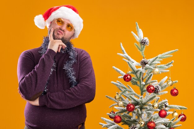 Homem pensativo de meia-idade com chapéu de Papai Noel e guirlanda de ouropel no pescoço e óculos perto da árvore de Natal decorada