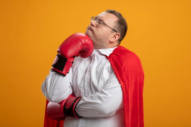 Homem pensativo super-herói eslavo adulto com capa vermelha usando óculos e luvas box, colocando a mão no queixo com os olhos fechados, isolado em um fundo laranja com espaço de cópia