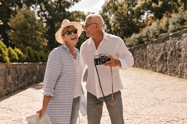 Foto grátis homem positivo com cabelos grisalhos em uma camisa clara e jeans com câmera rindo com uma senhora loira de chapéu, óculos escuros e camisa azul listrada no parque.