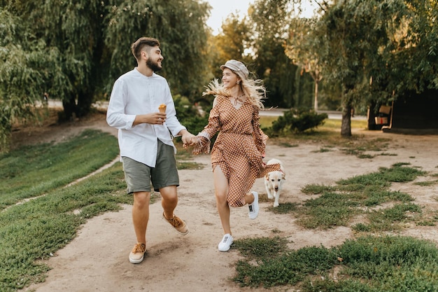 Homem positivo com sorvete está segurando a mão de uma mulher sorridente de vestido marrom casal romântico andando com grande labrador no parque