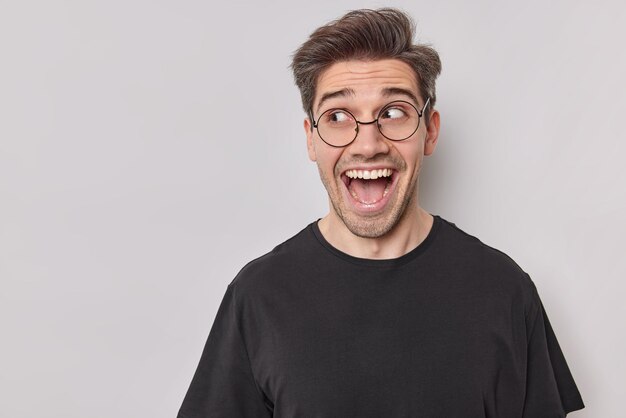 Homem positivo espantado parece feliz com a boca aberta e percebe que algo muito engraçado e surpreendente usa óculos redondos e camiseta preta isolada sobre fundo branco. Conceito de emoções