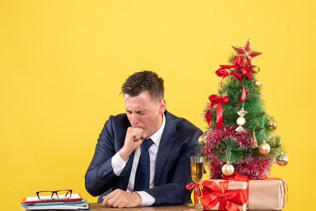 homem preocupado, fechando os olhos, sentado à mesa perto da árvore de natal e presentes em amarelo