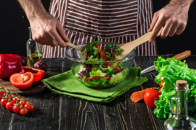 Homem preparando salada com legumes frescos em uma mesa de madeira. Cozinhar comida saborosa e saudável. Em fundo preto. Comida vegetariana, conceito saudável ou culinário.