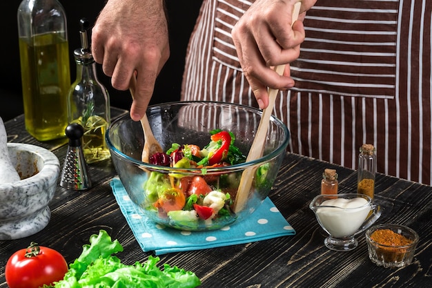 Homem preparando salada com legumes frescos em uma mesa de madeira. Cozinhar comida saborosa e saudável. Em fundo preto. Comida vegetariana, conceito saudável ou culinário.