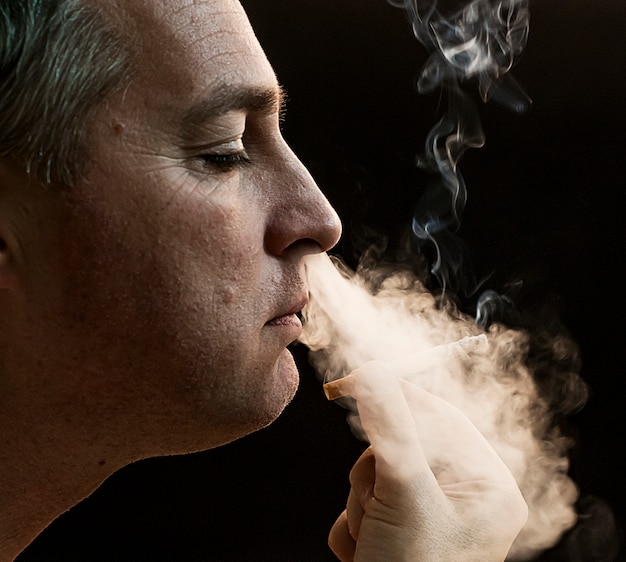 Homem que fuma cigarro no fundo preto, homem novo bonito que fuma cigarro, homem misterioso com charuto e fumo isolado no fundo preto