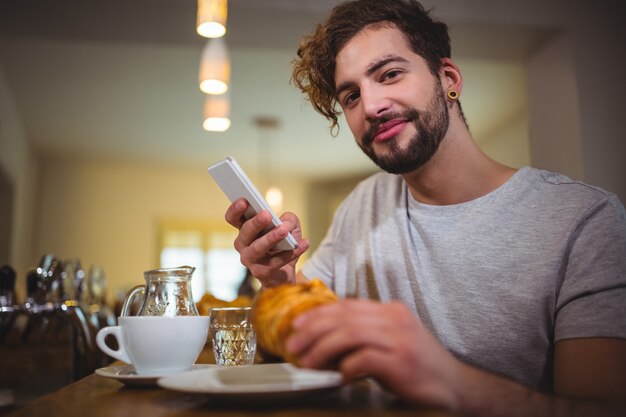 Homem que usa o telefone móvel ao ter croissant no cafÃ ©