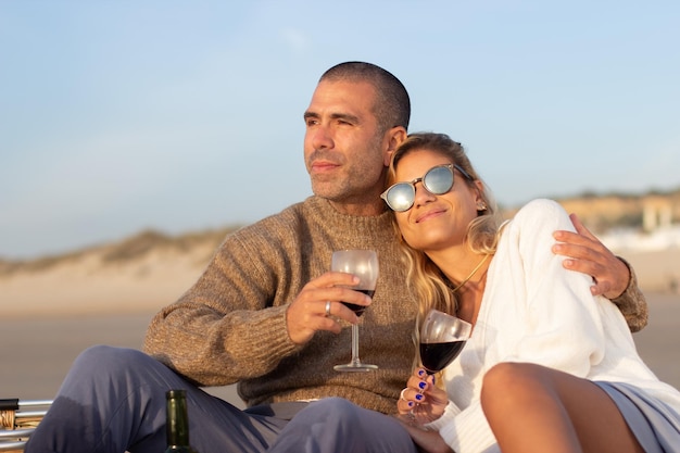Homem relaxado e mulher bebendo vinho na praia. casal fazendo piquenique, mulher encostada no ombro do marido. piquenique, bebida, conceito de relaxamento