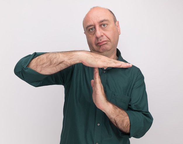 Homem rígido de meia-idade vestindo camiseta verde e mostrando gesto de tempo limite isolado na parede branca
