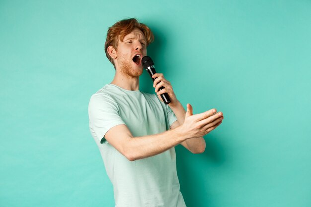 Homem ruivo apaixonado em uma camiseta cantando serenata com microfone, olhando de lado para o karaokê e gesticulando, em pé sobre o fundo de hortelã.