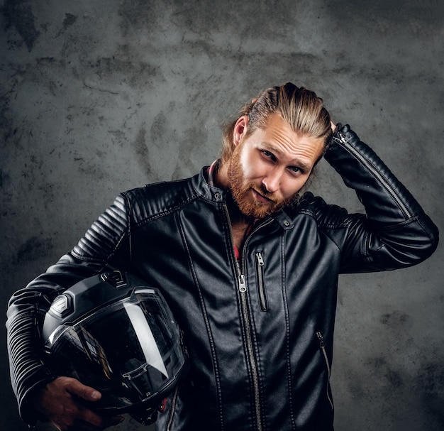 Foto grátis homem ruivo barbudo positivo na jaqueta de couro detém capacete de motocicleta em fundo cinza.