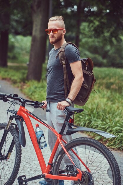 Homem ruivo bonito com um corte de cabelo elegante e barba vestida com roupas esportivas e óculos de sol caminha no parque com uma bicicleta e mochila.