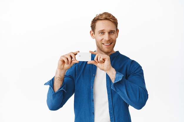 Homem ruivo bonito mostrando seu cartão de crédito e sorrindo satisfeito, anúncio de banco, compras ou promoção de descontos especiais, em pé sobre uma parede branca