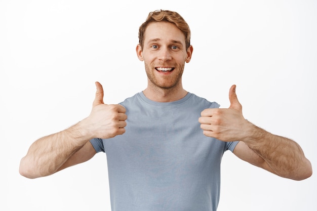 Homem ruivo bonito sorridente mostrando os polegares em aprovação, curtindo e concordando, elogiando e recomendando a oferta promocional, satisfeito com alguma coisa, em pé sobre uma parede branca