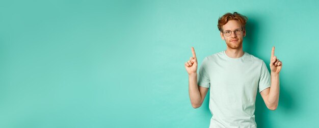 Homem ruivo cético e sem graça de óculos e camiseta apontando os dedos para cima e mostrando algo u