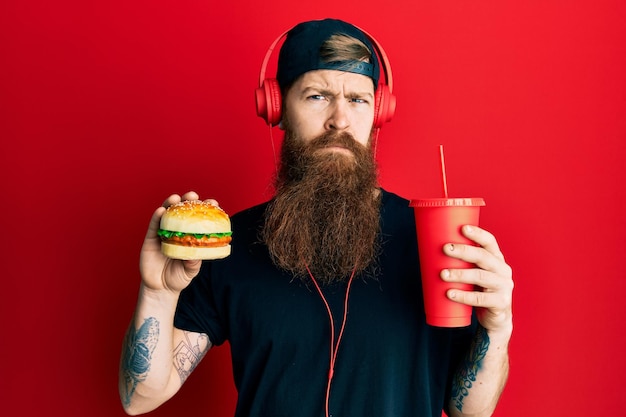 Homem ruivo com barba longa comendo um saboroso hambúrguer clássico e bebendo refrigerante cético e nervoso, carrancudo chateado por causa do problema. pessoa negativa.