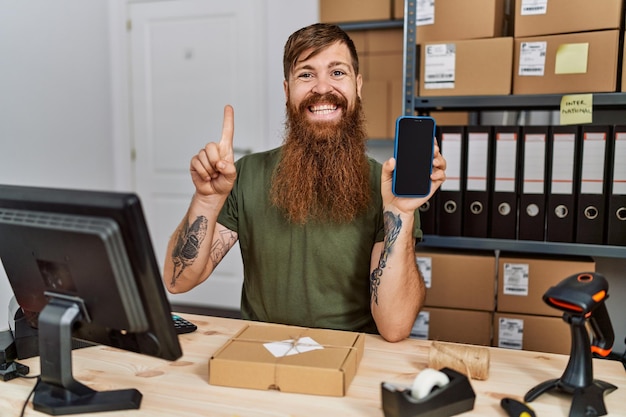 Homem ruivo com barba longa trabalhando em pequenas empresas segurando smartphone sorrindo com uma ideia ou pergunta apontando o dedo com cara feliz número um