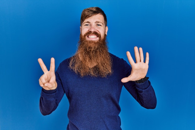 Homem ruivo com barba longa vestindo suéter azul casual sobre fundo azul mostrando e apontando para cima com os dedos número sete enquanto sorria confiante e feliz.