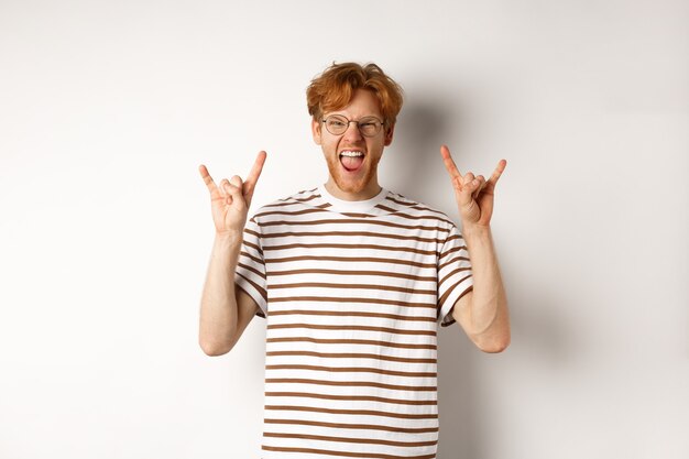 Homem ruivo engraçado e feliz se divertindo, mostrando o chifre de rock-n-roll e enfiando a língua, aproveitando a festa, em pé sobre um fundo branco.