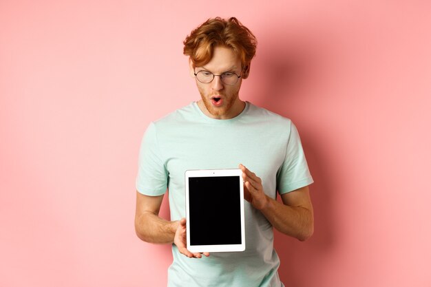 Homem ruivo impressionado de óculos, mostrando a tela do tablet digital em branco e olhando maravilhado ...