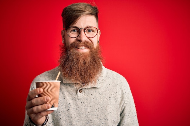 Homem ruivo irlandês com barba bebendo um café para viagem em um copo de papel sobre fundo vermelho com um rosto feliz de pé e sorrindo com um sorriso confiante mostrando os dentes